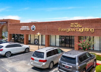 A brick shopping plaza called Heritage Plaza houses an animal hospital and EverGlow Lighting; parked cars, flowering shrubs, and a bright blue sky create a sunny, welcoming scene.